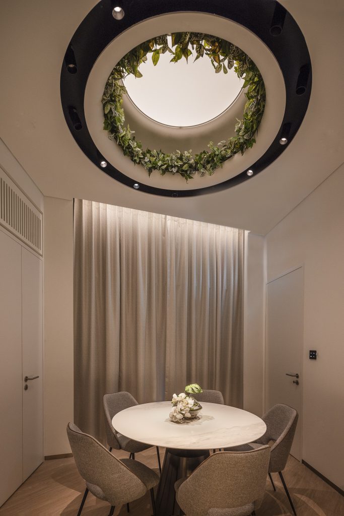 A small table and some chairs in a room with a mock skylight lined with greenery